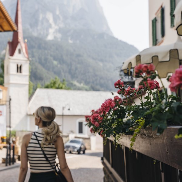 Sensoria Dolomites: Wellnesshotel in den Dolomiten Frau spaziert in Bergdorf mit Blumen und Kirche im Hintergrund