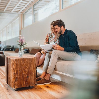 Naturhotel in Südtirol. Luxus für die Seele. Paar sitzt auf Sofa und liest Buch im modernen Raum mit Holztisch und Vase