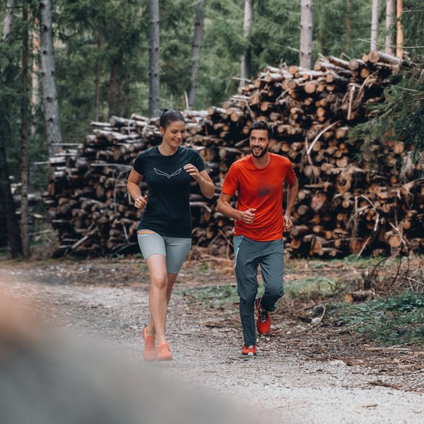 Active holidays in South Tyrol – ready for pure nature? Man and woman running on forest path next to stacked logs