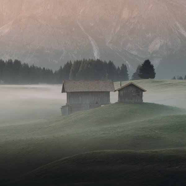 Sustainability at Sensoria Dolomites Foggy landscape with two wooden huts on a green hill and mountains in the background