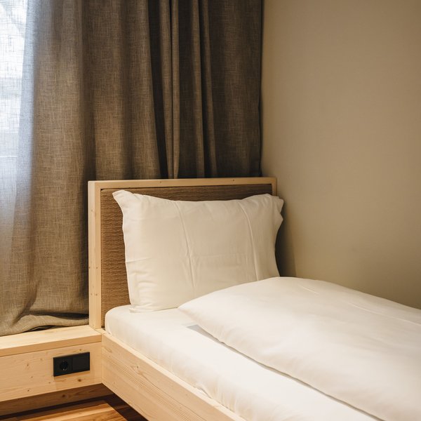 Romantischer Kurzurlaub für Verliebte – Sensoria Dolomites Einzelbett mit weißer Bettwäsche vor braunem Vorhang und Holzboden