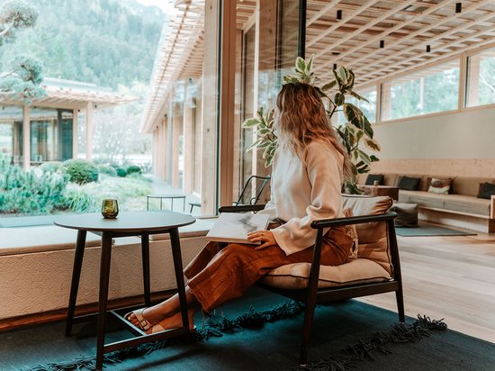 Größte Hochalm Europas – ein Kraftplatz mit Seele Frau sitzt mit Buch in modernem Raum und blickt aus großem Fenster auf Garten