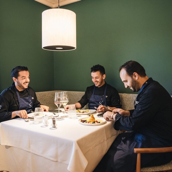 Genussurlaub in Südtirol – Sensoria Dolomites Drei Männer in Küchenuniformen sitzen an einem gedeckten Tisch und lachen