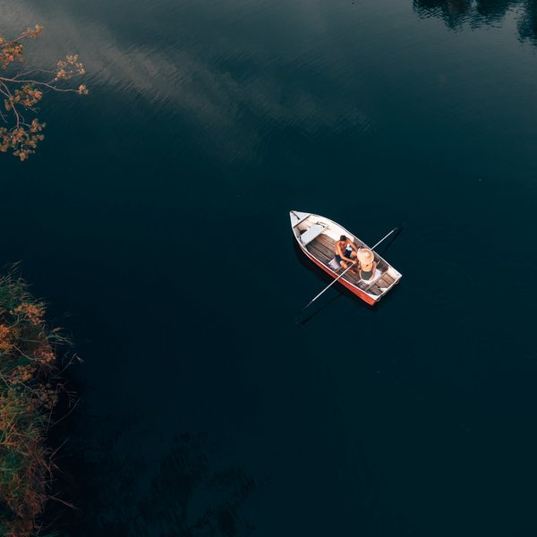 Meteo all’Alpe di Siusi? Soleggiato! Due persone sedute su una barca su acqua calma vicino alla riva con vegetazione