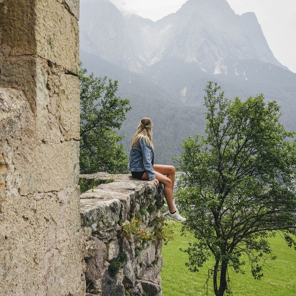 Sensoria Dolomites: Wellnesshotel in den Dolomiten Frau sitzt auf alter Steinmauer mit Blick auf Berge und grüne Landschaft