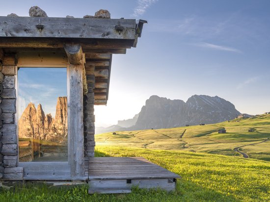 A summer holiday in the mountains Mountain cabin reflecting Dolomites in window with green meadow in sunlight