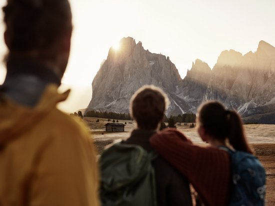 Sensoria Dolomites: your hotel in the Italian Alps Three people looking at the mountains at sunset in a field with a cabin