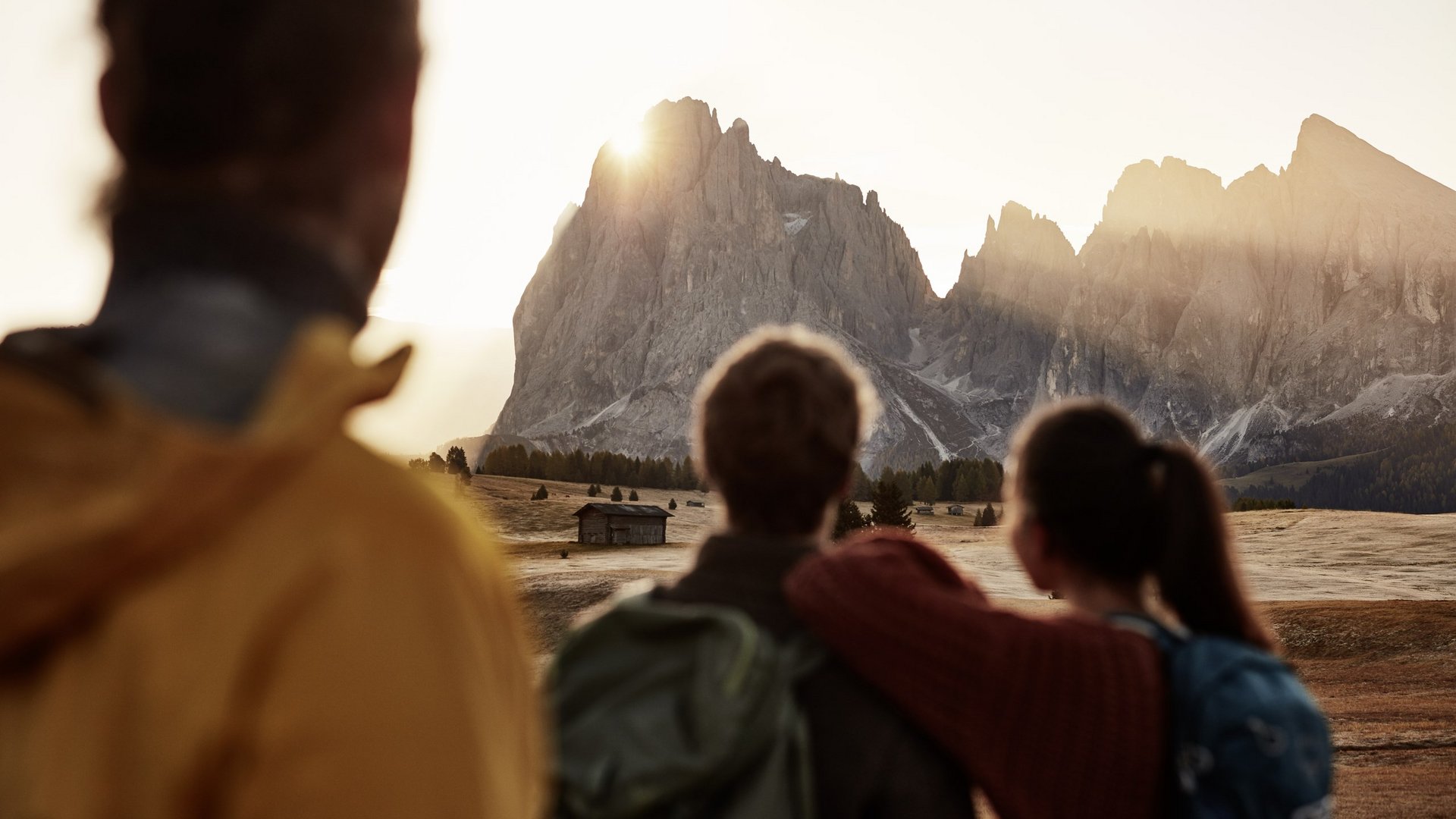 Alpe di Siusi: cosa fare in estate al Sensoria Dolomites Tre persone guardano le montagne al tramonto in una radura con una baita