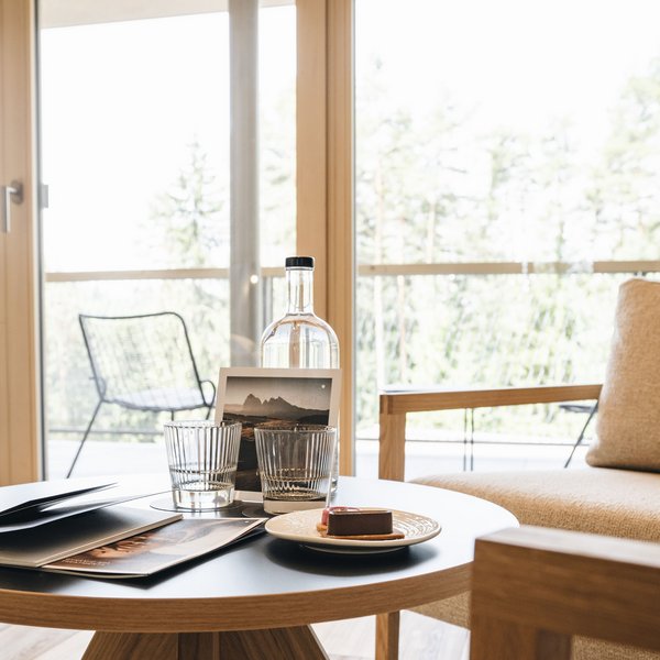 Romantischer Kurzurlaub für Verliebte – Sensoria Dolomites Moderner Wohnzimmer-Tisch mit Wasserflasche, zwei Gläsern und Dessert vor Balkon
