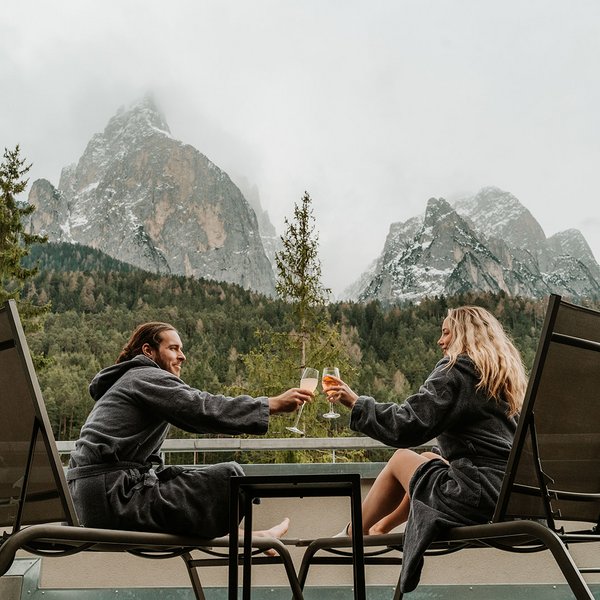 Sensoria Dolomites: Wellnesshotel in den Dolomiten Paar in Bademänteln stößt mit Getränken auf Terrasse mit Bergblick an