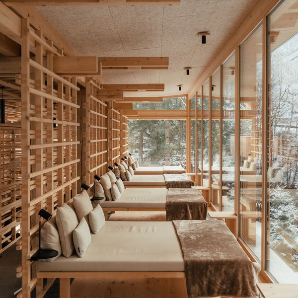 Hotel in mezzo alla natura: area spa del Sensoria Dolomites Lettini in legno chiaro con cuscini in una stanza con ampie finestre e vista invernale