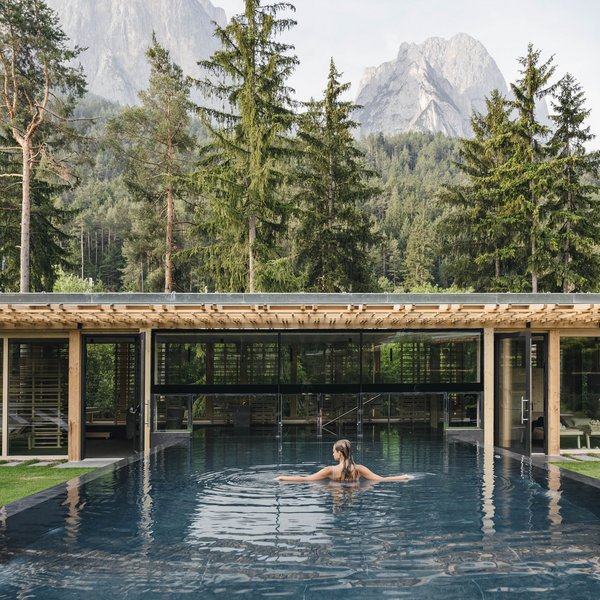 Sensoria Dolomites: Wellnesshotel in den Dolomiten Frau im Pool mit Blick auf bewaldete Berge und moderne Gebäude
