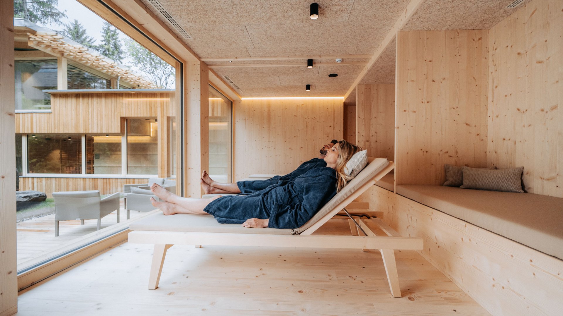 From hiking on Alpe di Siusi/Seiser Alm to the spa area Couple relaxing in blue robes on wooden loungers in modern spa with window view