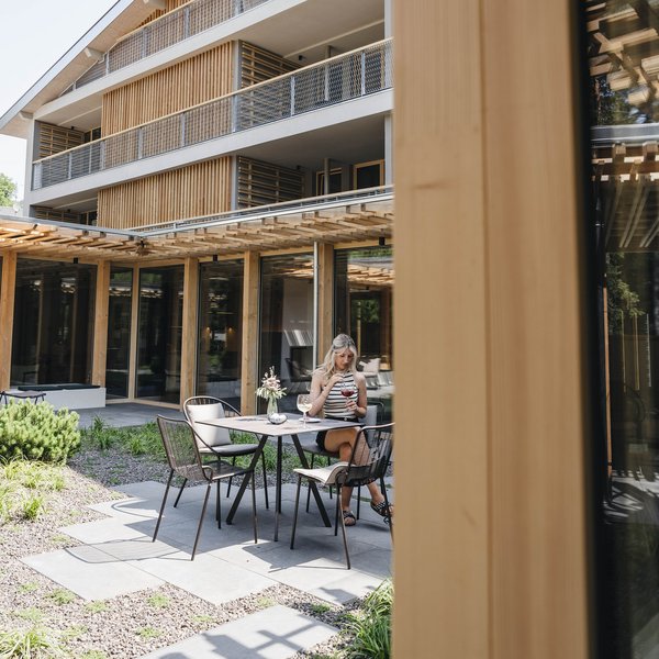 Sensoria Dolomites: Wellnesshotel in den Dolomiten Frau sitzt draußen an Tisch mit Getränken vor modernem Gebäude