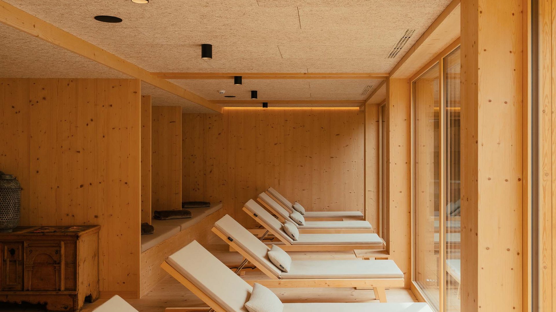 Das Badehaus im Hotel mit Pool in den Dolomiten Entspannungsbereich mit Liegestühlen in einem Holzinterieur mit großen Fenstern