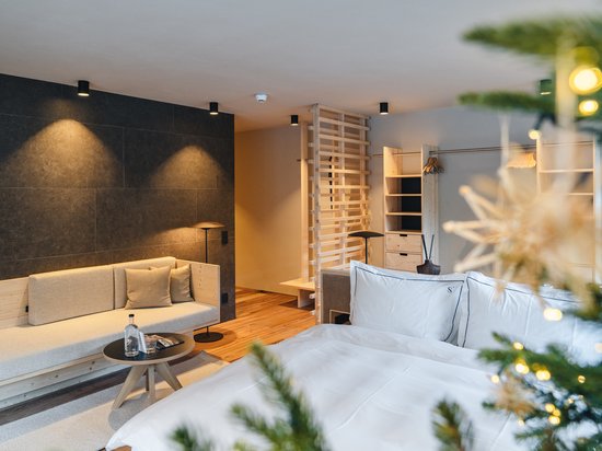 Sensoria Dolomites – Urlaub in Seis am Schlern Modernes Hotelzimmer mit großem Bett, Sofa und warmem Licht, eingerahmt von Tannenzweigen