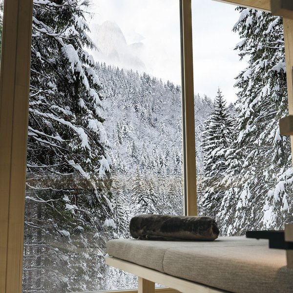 Hotel in mezzo alla natura: area spa del Sensoria Dolomites Vista sulla foresta innevata attraverso finestra vicino a lettino in legno con coperta