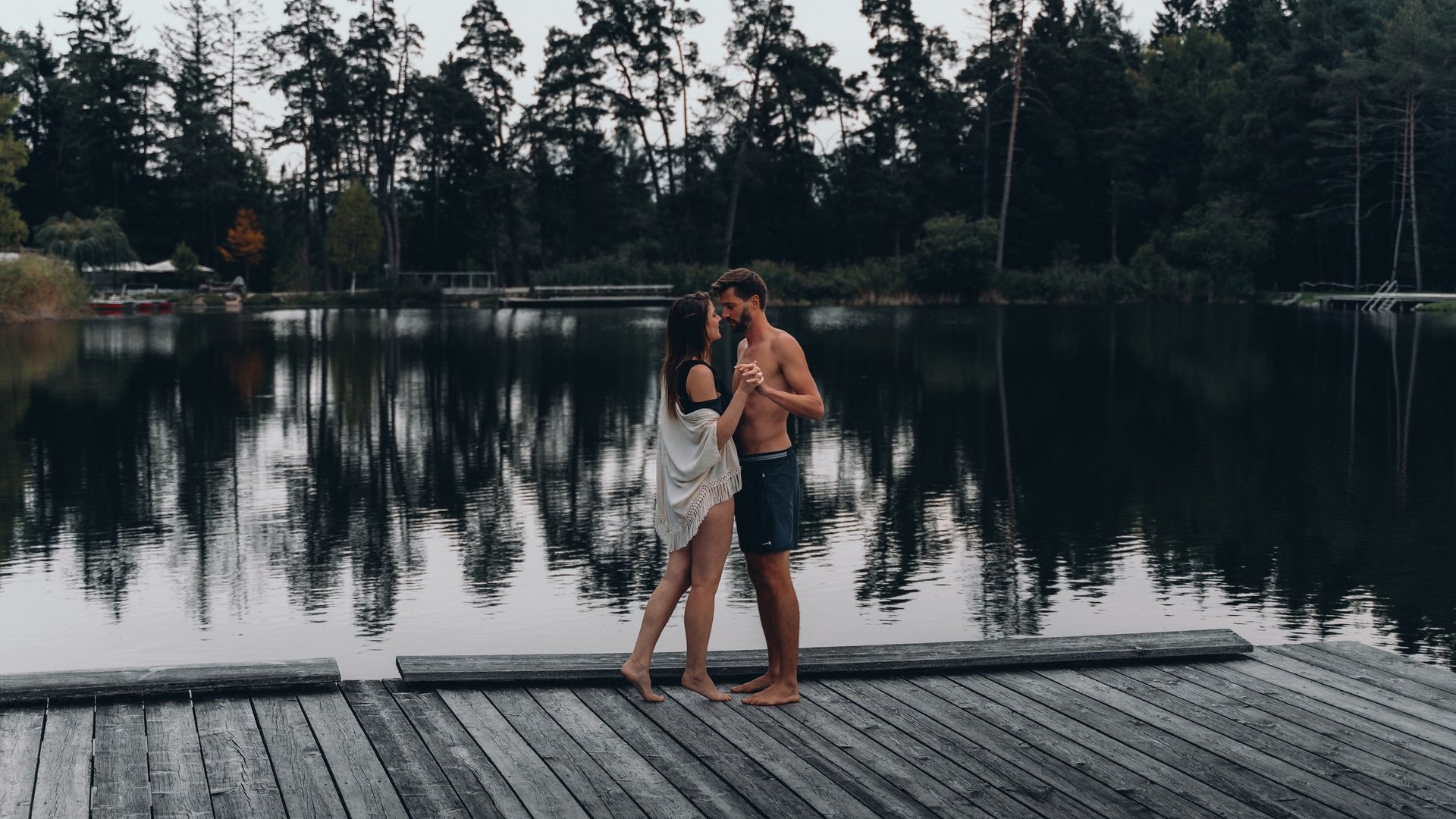 Kostbare Augenblicke in Ihrem Romantikhotel in den Dolomiten Paar tanzt am Steg vor ruhigem See mit Wald im Hintergrund
