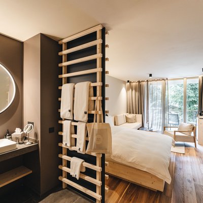 Naturhotel in Südtirol. Luxus für die Seele. Modernes Hotelzimmer mit Holzfußboden, Bett, Sofa und beleuchtetem Badezimmer