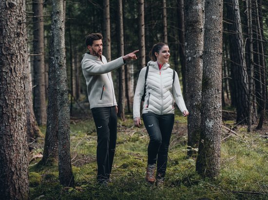 Hotel a Siusi allo Sciliar? Sensoria Dolomites! Coppia che cammina nel bosco, uomo indica qualcosa, entrambi vestiti outdoor