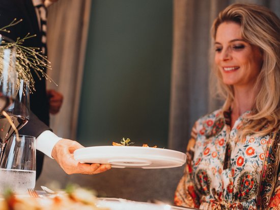 Kostbare Augenblicke in Ihrem Romantikhotel in den Dolomiten Kellner serviert einem lächelnden Frau in gemustertem Kleid ein Gericht