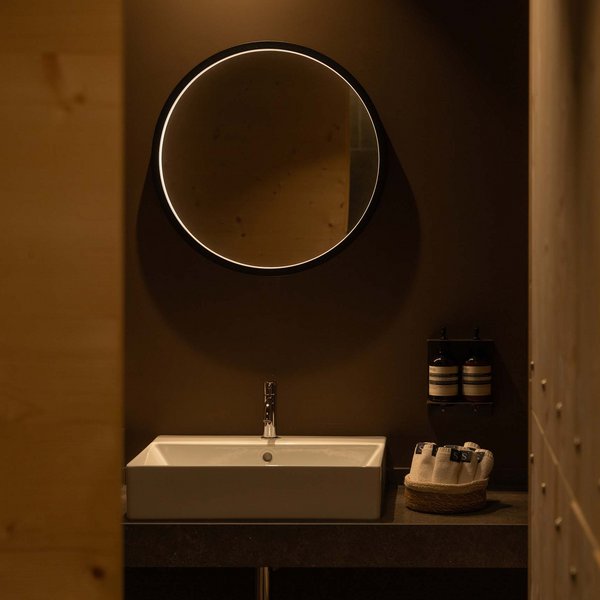 Romantischer Kurzurlaub für Verliebte – Sensoria Dolomites Beleuchteter runder Spiegel über Waschbecken und Handtüchern im dunklen Badezimmer