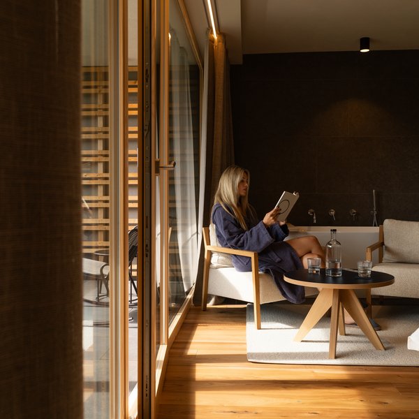 Sensoria Dolomites: Wellnesshotel in den Dolomiten Frau entspannt im Bademantel im modernen Wohnzimmer und liest Buch