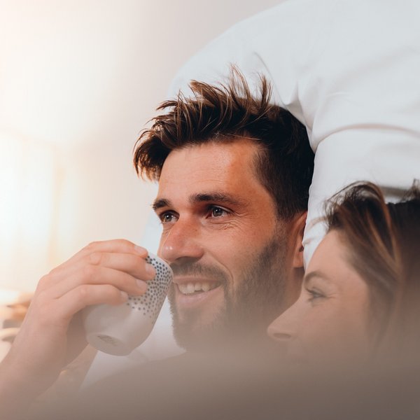 Romantischer Kurzurlaub für Verliebte – Sensoria Dolomites Paar entspannt im Bett, Mann trinkt aus einer Tasse