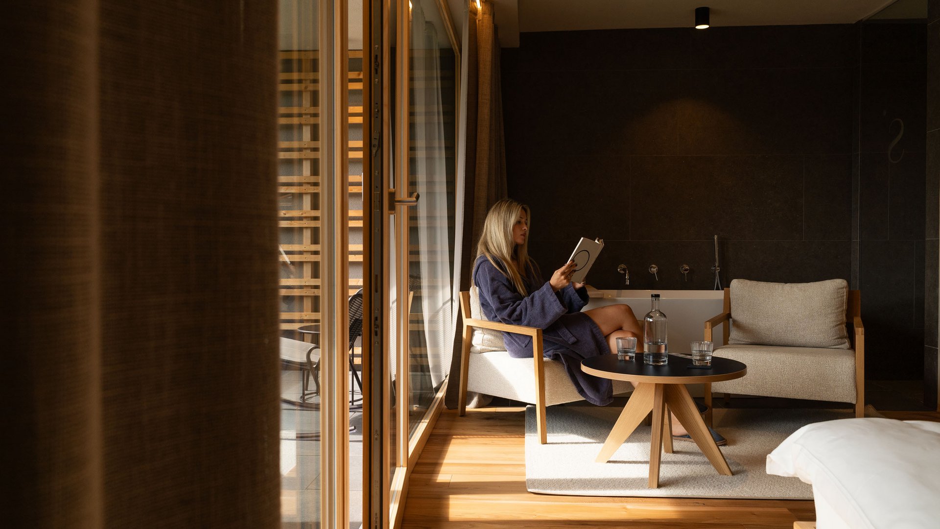 Sensoria Dolomites: Wellnesshotel in den Dolomiten Frau liest in Bademantel in einem modernen Hotelzimmer mit Tageslicht