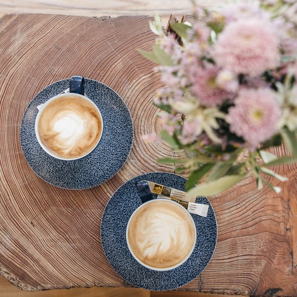 Sensoria Dolomites: Babymoon in Südtirol Zwei Tassen Latte Macchiato mit Milchschaum auf Holztisch mit Blumen