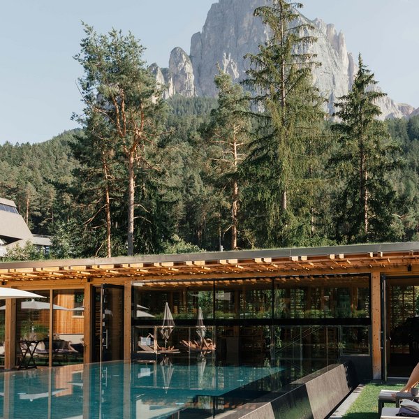 From hiking on Alpe di Siusi/Seiser Alm to the spa area Modern pool with glass building in front of forested mountain in sunny landscape