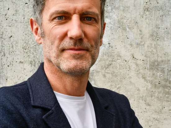 My Olympic Mindset Retreat 2026 Uomo con capelli grigi e giacca nera davanti a muro di cemento grigio